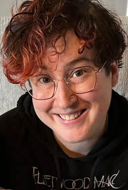 Picture of white person with round wireframe glasses, red and brown curly hair, wearing a black sweater