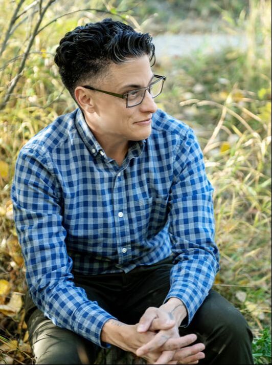 Image of Nathan, a light-skinned racialized person with short cropped hair, bold squared glasses, and a blue checkered collared shirt, sitting with hands clasped, looking off-camera,  in front of tall grasses and a gravel path.